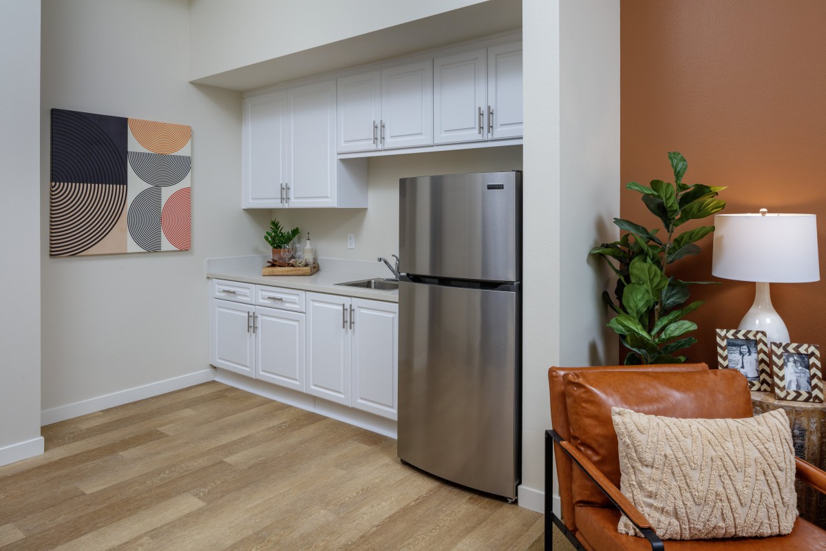A modern apartment kitchen with white cabinets, stainless steel fridge, and wood flooring.