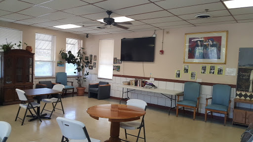 A cozy community room with a circular table, chairs, a television on the wall, and decorative plants.