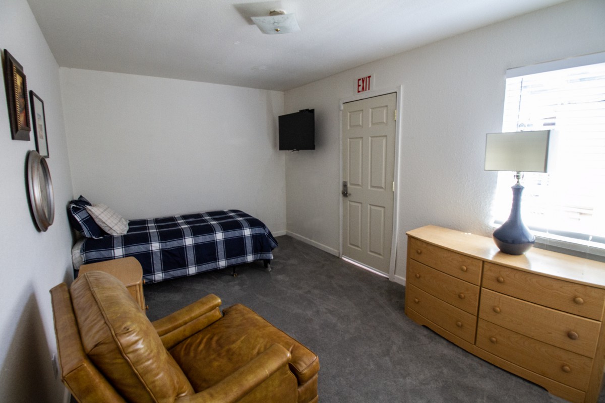A minimalist bedroom with a single bed, armchair, dresser, and wall-mounted television.