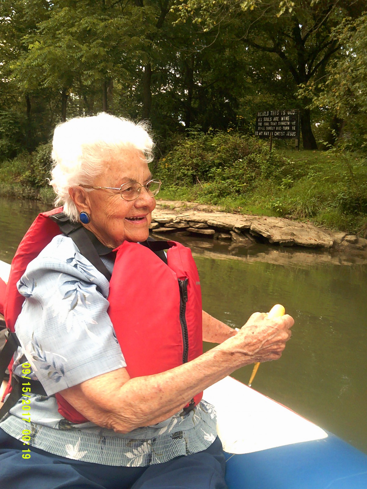 An elderly woman wearing a life jacket enjoys a boat ride on a peaceful river.