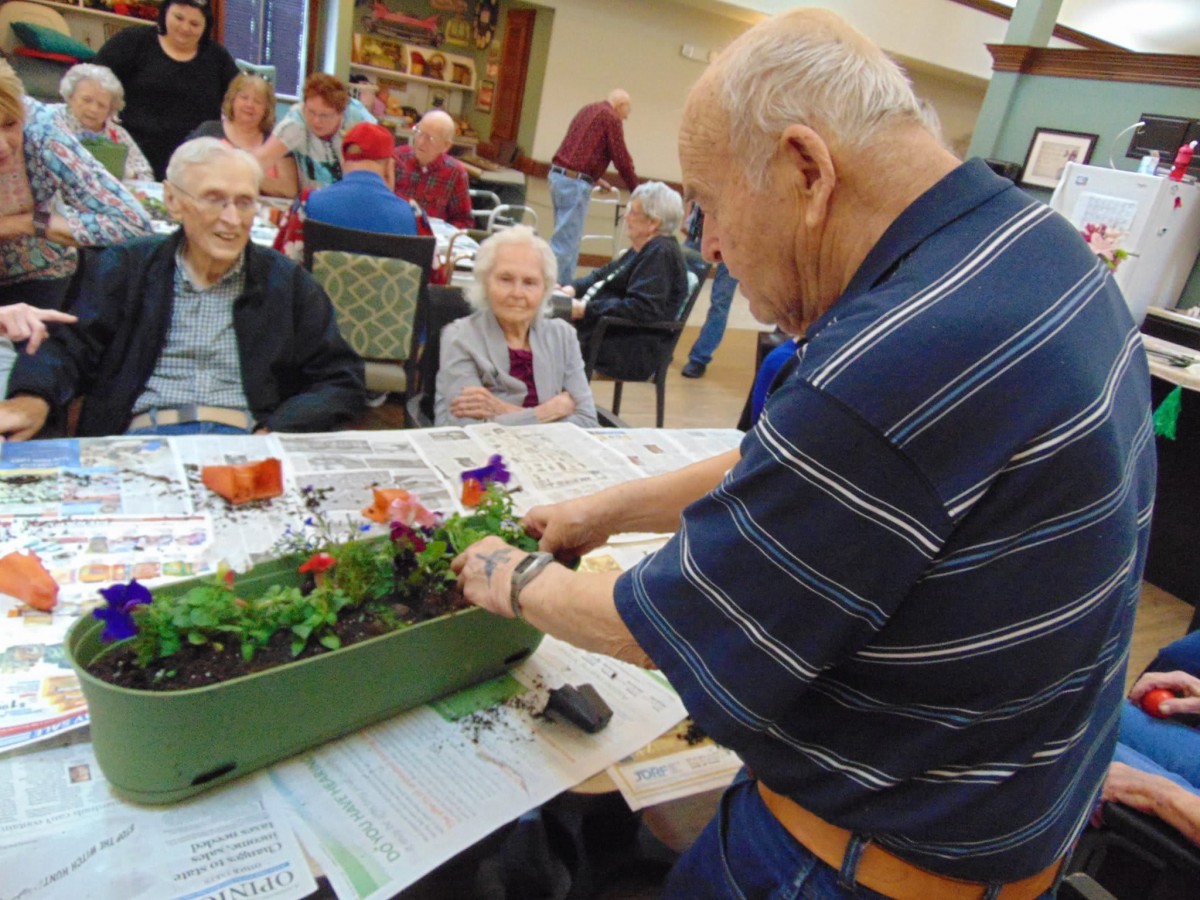 An elderly man tends to a planter while seated seniors watch in a community center setting.