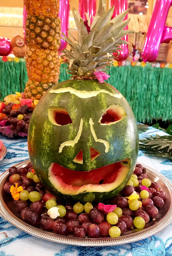 A creatively carved watermelon with a playful face, topped with a pineapple and surrounded by an assortment of fruits.