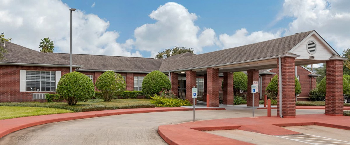 A brick building with a covered entrance and landscaped gardens under a blue, cloudy sky.