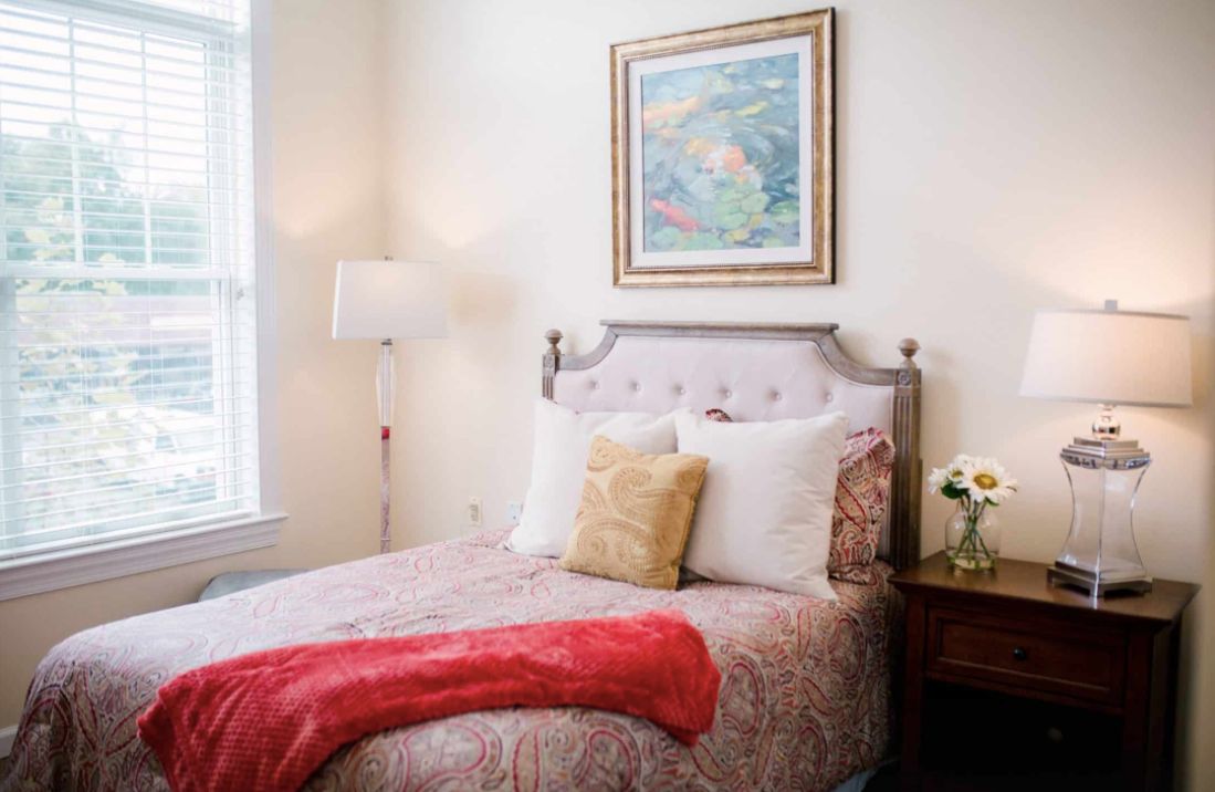 A cozy bedroom with a neatly made bed, framed painting above the headboard, and a floor lamp beside the bed.