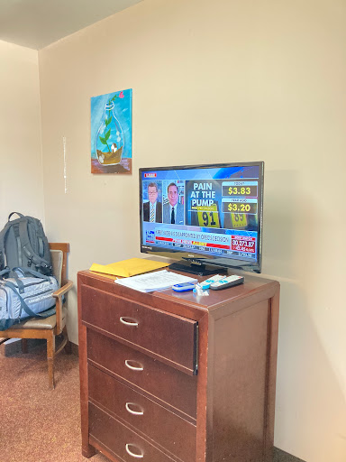 A cozy sitting area with a television displaying news, next to a wooden dresser and wall art.