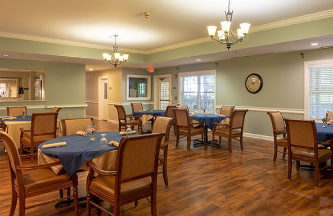 A cozy dining room with neatly arranged tables and chairs on a hardwood floor, illuminated by elegant lighting fixtures.