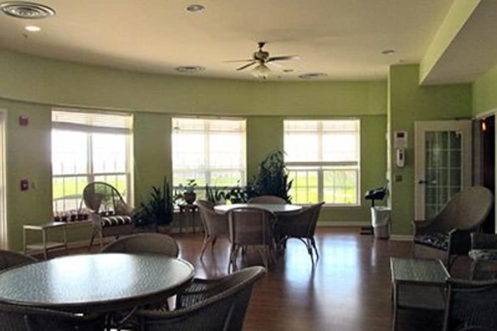 A spacious room with tables and chairs, featuring large windows and plants, under a ceiling fan.