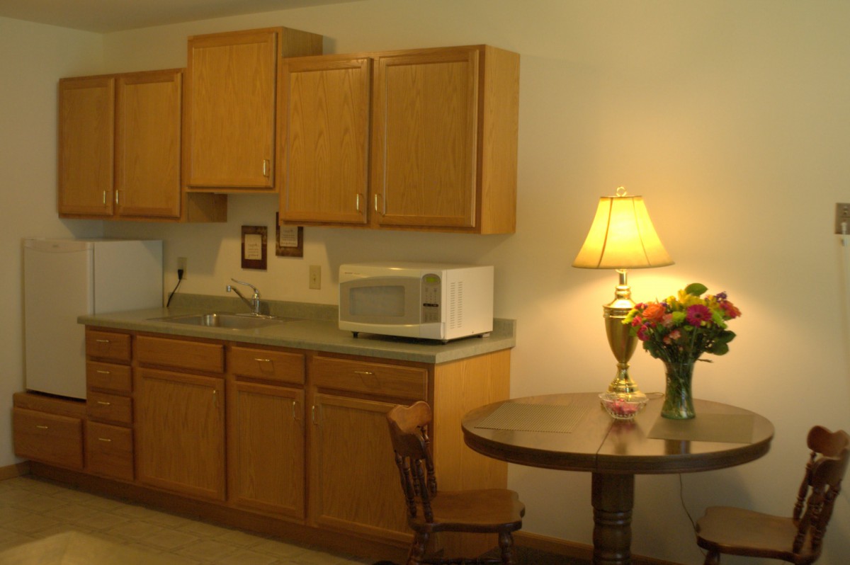 A cozy kitchen area with wooden cabinets, a microwave, and a small round table with a lamp and flowers.
