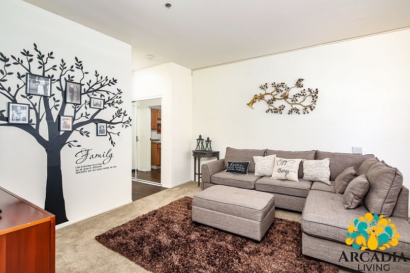 A cozy living room with a gray sofa, wall art featuring a family tree decal, and decorative cushions on a plush rug.