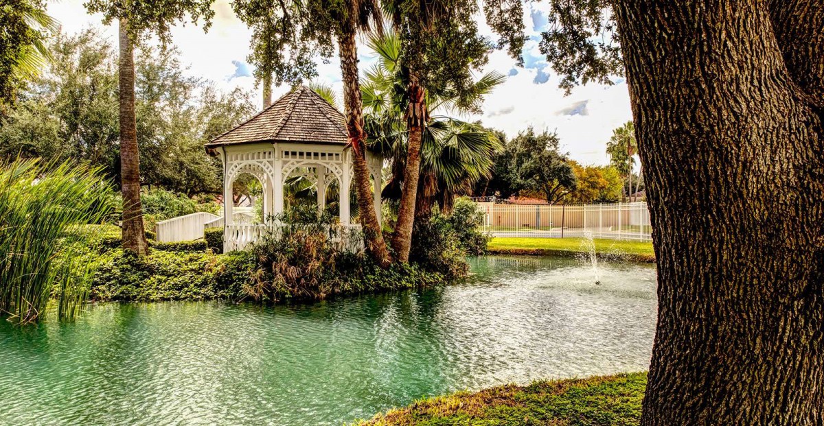 A tranquil garden scene featuring a gazebo by a pond with a fountain, surrounded by lush greenery.