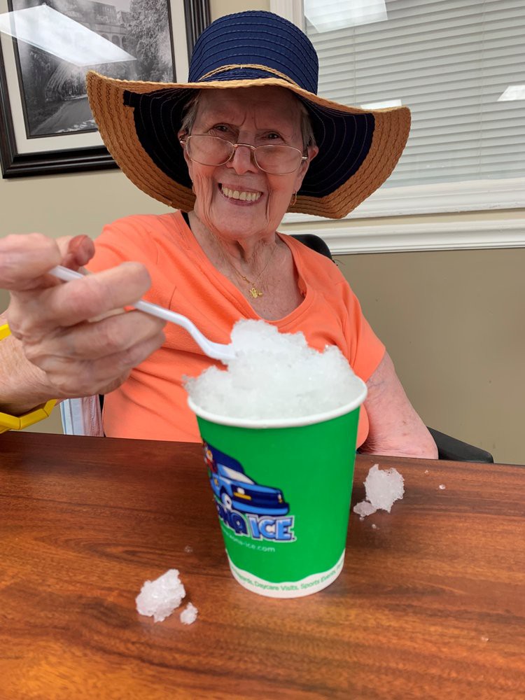 An elderly woman wearing a wide-brimmed hat and orange shirt enjoys a cup of shaved ice, smiling at the camera.