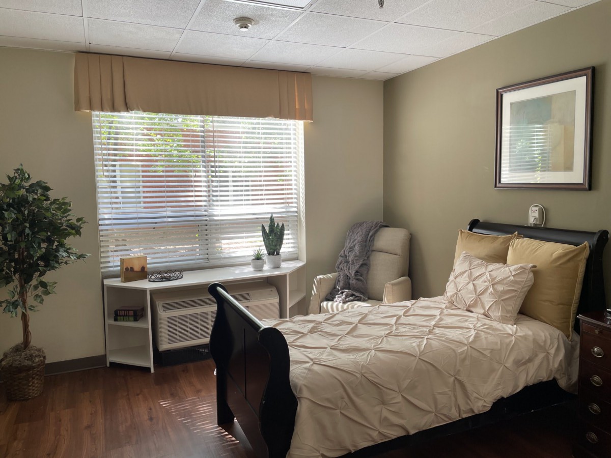 A cozy and neatly arranged bedroom with a single bed, armchair, and window with blinds.