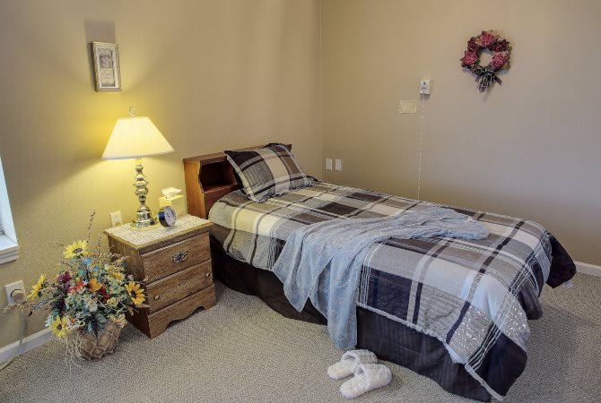 A cozy bedroom with a single bed and nightstand, featuring a lamp and slippers.