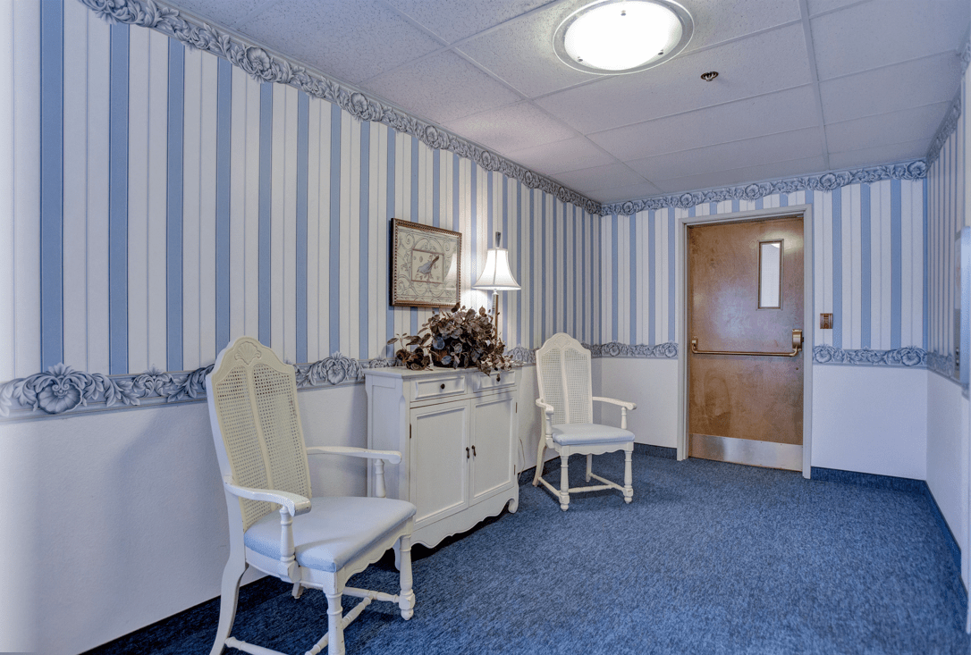 A cozy waiting area featuring two white chairs, a lamp on a dresser, and striped wallpaper.