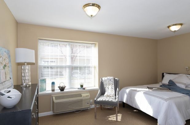 A minimalist bedroom with beige walls, a large window allowing natural light, and simple furnishings.