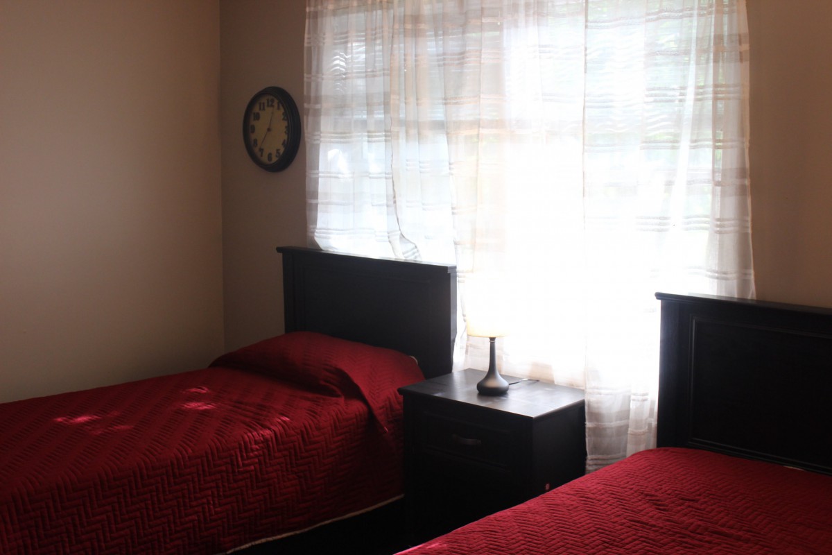 A cozy bedroom with two twin beds featuring red bedding, a window with sheer curtains, and a nightstand with a lamp.