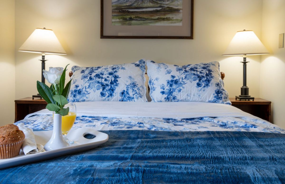 A cozy bedroom setup featuring a bed with blue and white floral pillows, a matching comforter, and a tray with a drink and plant on top. The space is warmly lit by lamps on either side.