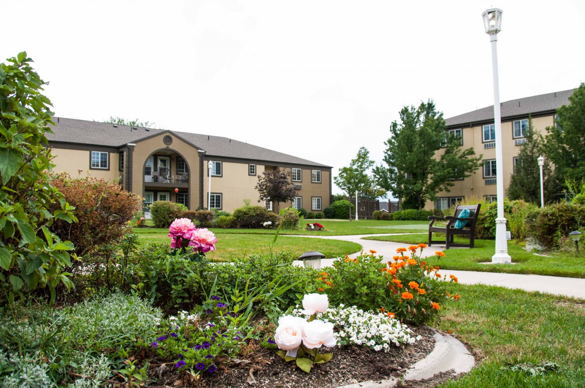 A view of apartment buildings with a landscaped garden featuring colorful flowers and a winding pathway under a cloudy sky.