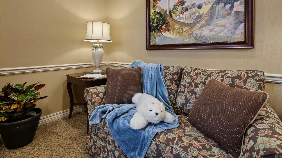 A cozy living room with a floral sofa, accented by a blue blanket and stuffed bear, set beside a lamp and painting.
