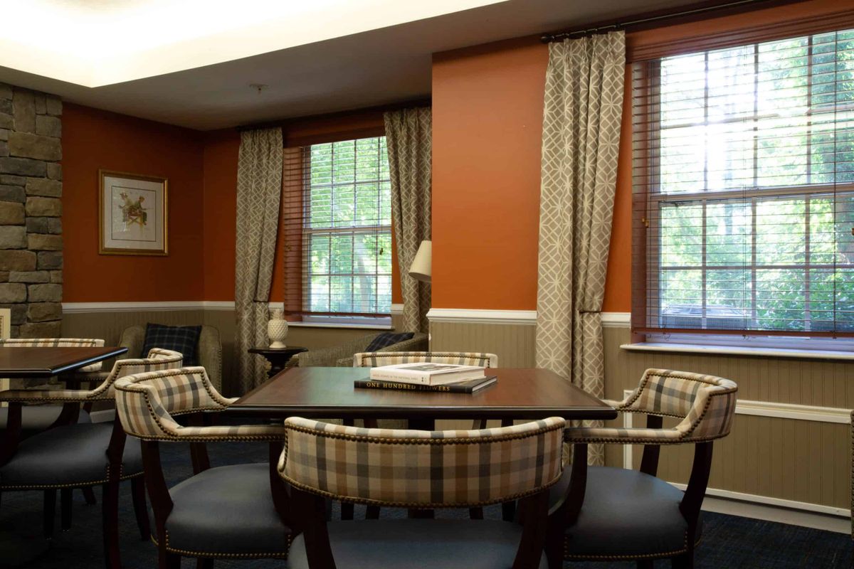 A cozy room with a wooden table and cushioned chairs, flanked by windows with patterned curtains.
