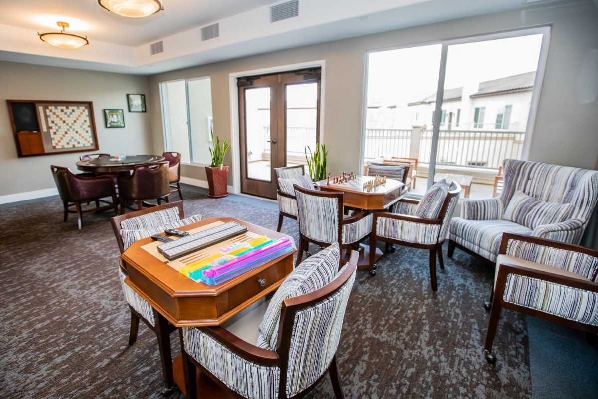 A well-lit indoor room featuring tables and chairs arranged for board games, illuminated by sunlight streaming through large windows.
