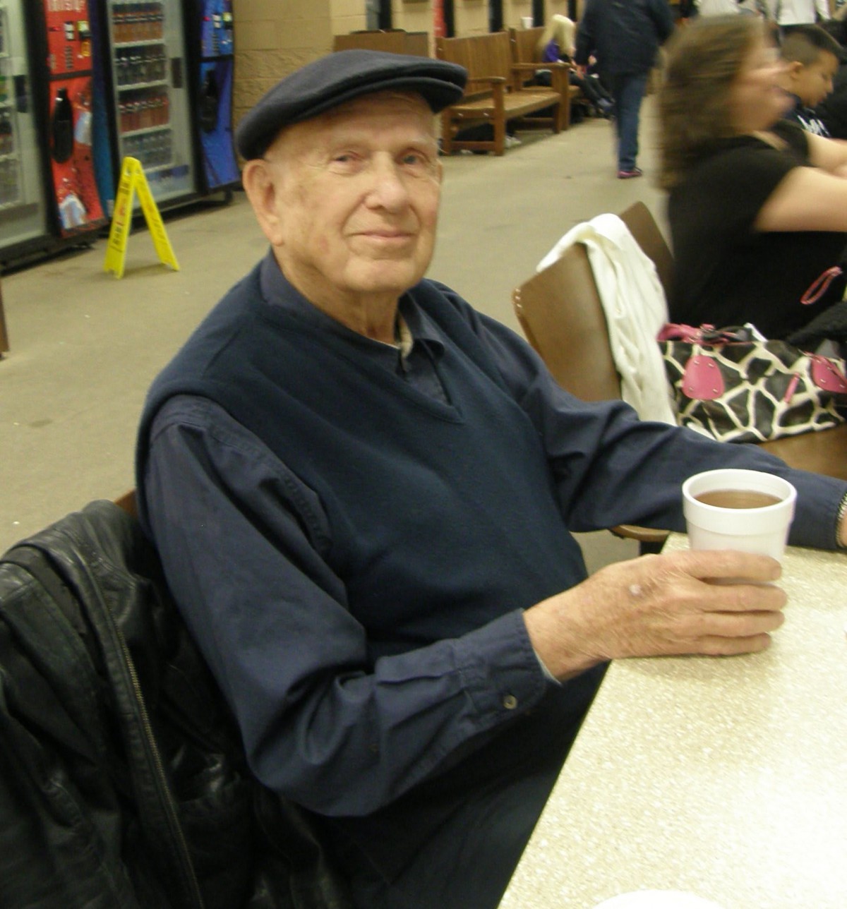 A man wearing a hat is sitting at a table holding a cup of coffee and smiling.