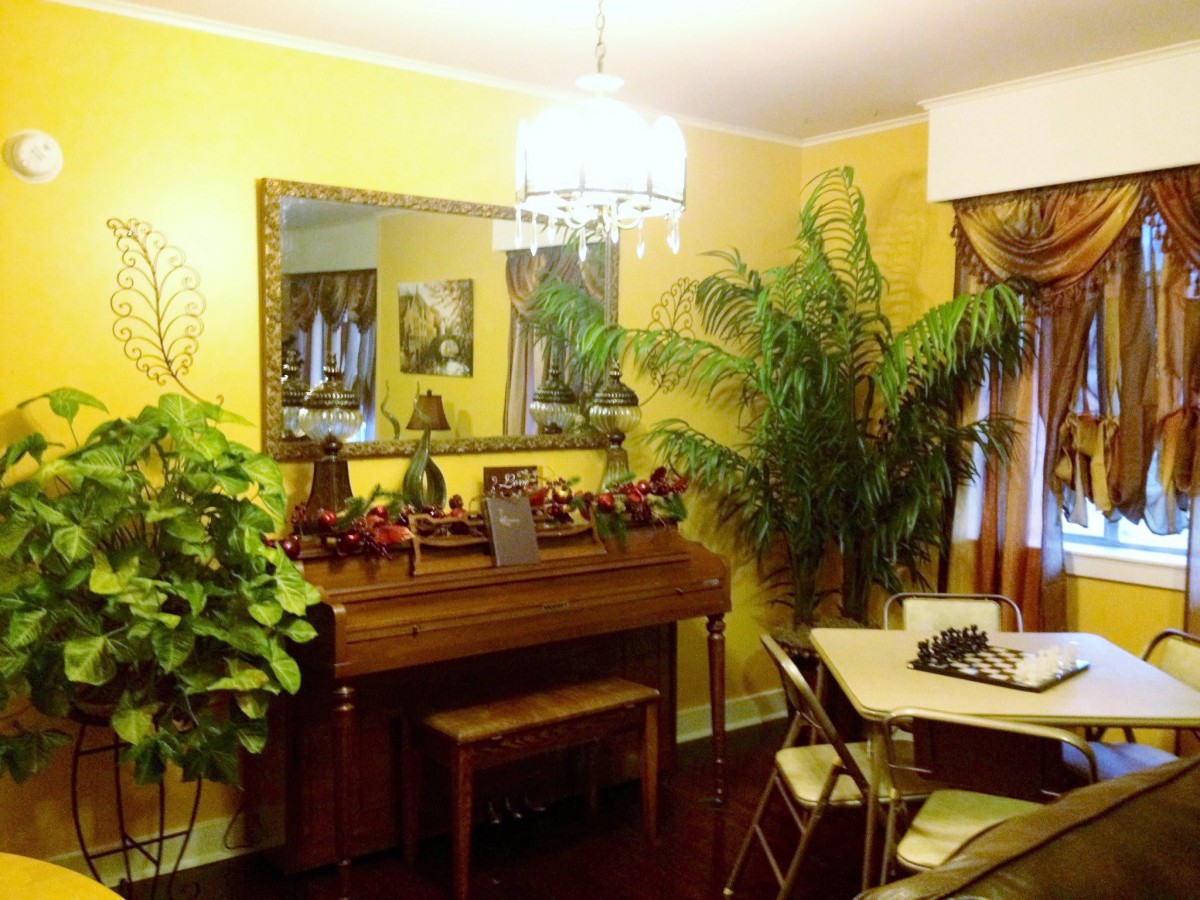 A cozy dining room featuring a piano, lush plants, and a chandelier against vibrant yellow walls.