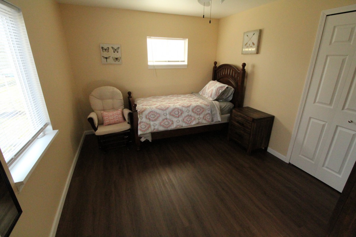 A minimalist bedroom featuring a single bed, a chair, and a nightstand on a dark hardwood floor.
