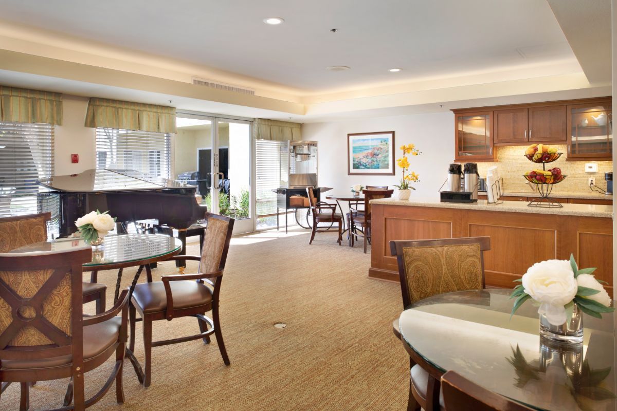 A cozy dining area with tables, chairs, a piano, and a coffee station under bright lighting.