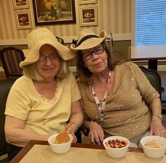 Two elderly women wearing hats and seated indoors, enjoying snacks.