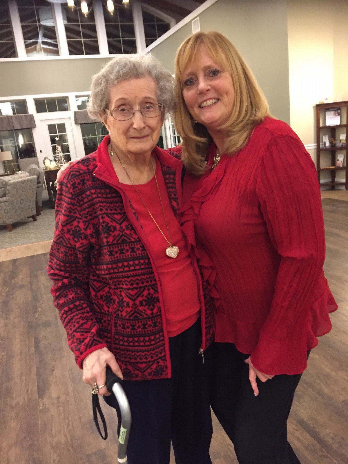 An elderly woman and a younger woman, both in red outfits, smiling indoors.