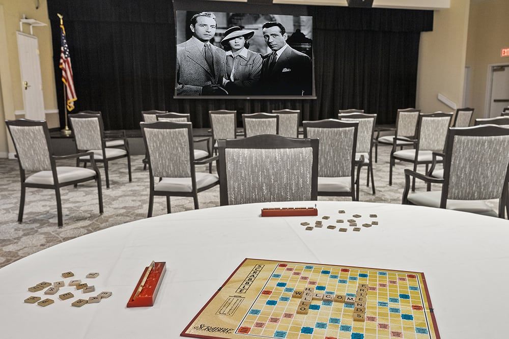 A Scrabble board is set up on a table in a room with chairs and a classic movie poster in the background.