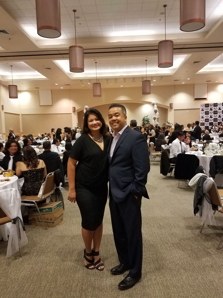 A couple dressed in formal attire poses in a crowded dining hall during an event.
