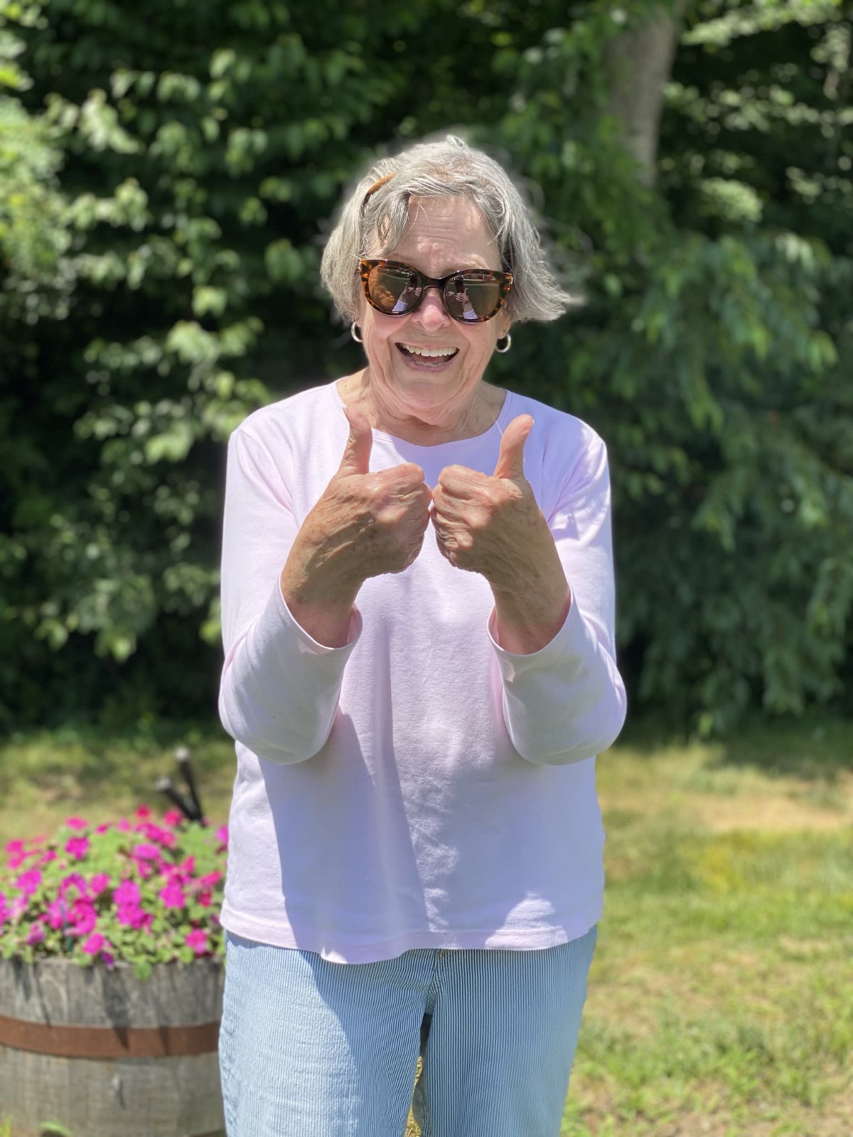 A woman wearing sunglasses gestures cheerfully outdoors on a sunny day.