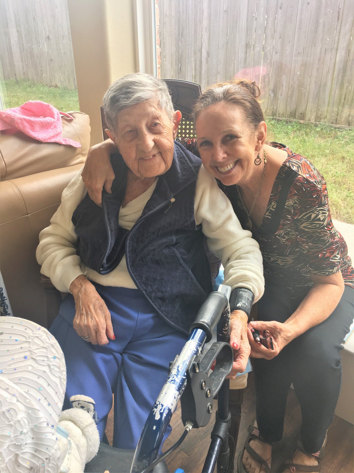 An elderly person sitting in a wheelchair with a younger woman hugging and smiling beside them outdoors.