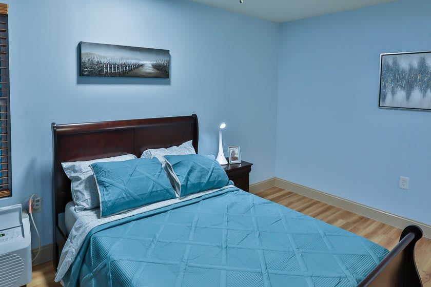 A cozy bedroom with blue walls, featuring a bed with blue bedding, a nightstand lamp, and framed artwork.