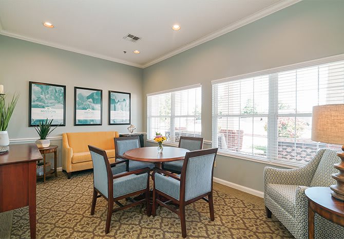 A cozy living room with a round table surrounded by chairs, large windows letting in natural light, and artwork adorning the walls.