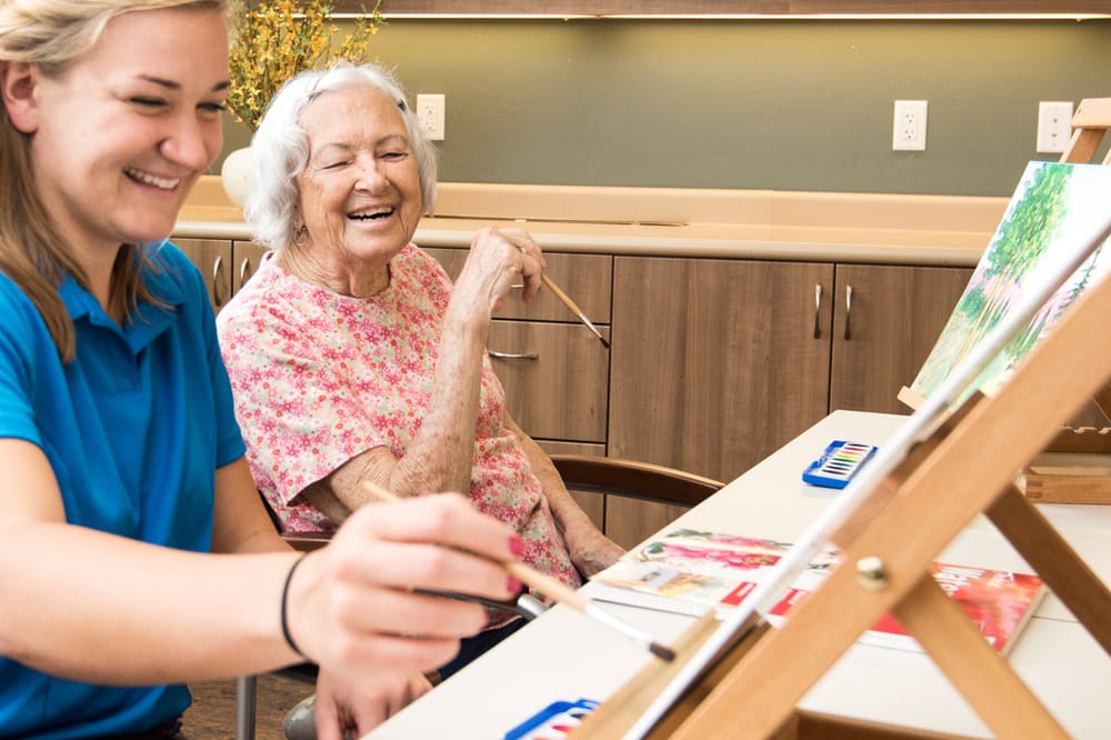 An elderly woman smiling as she engages in a painting activity indoors.