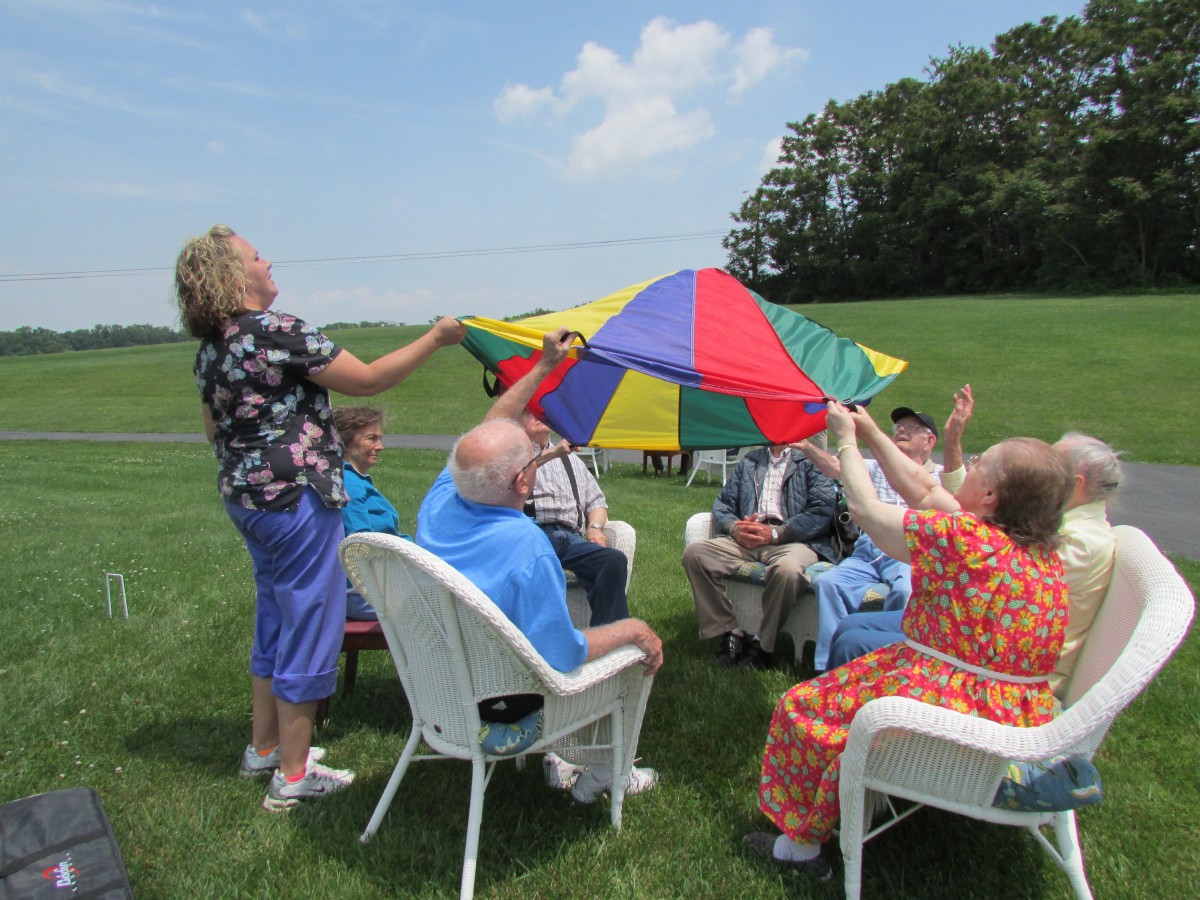 A group of adults sitting in chairs outdoors, participating in a parachute activity on a grassy field.