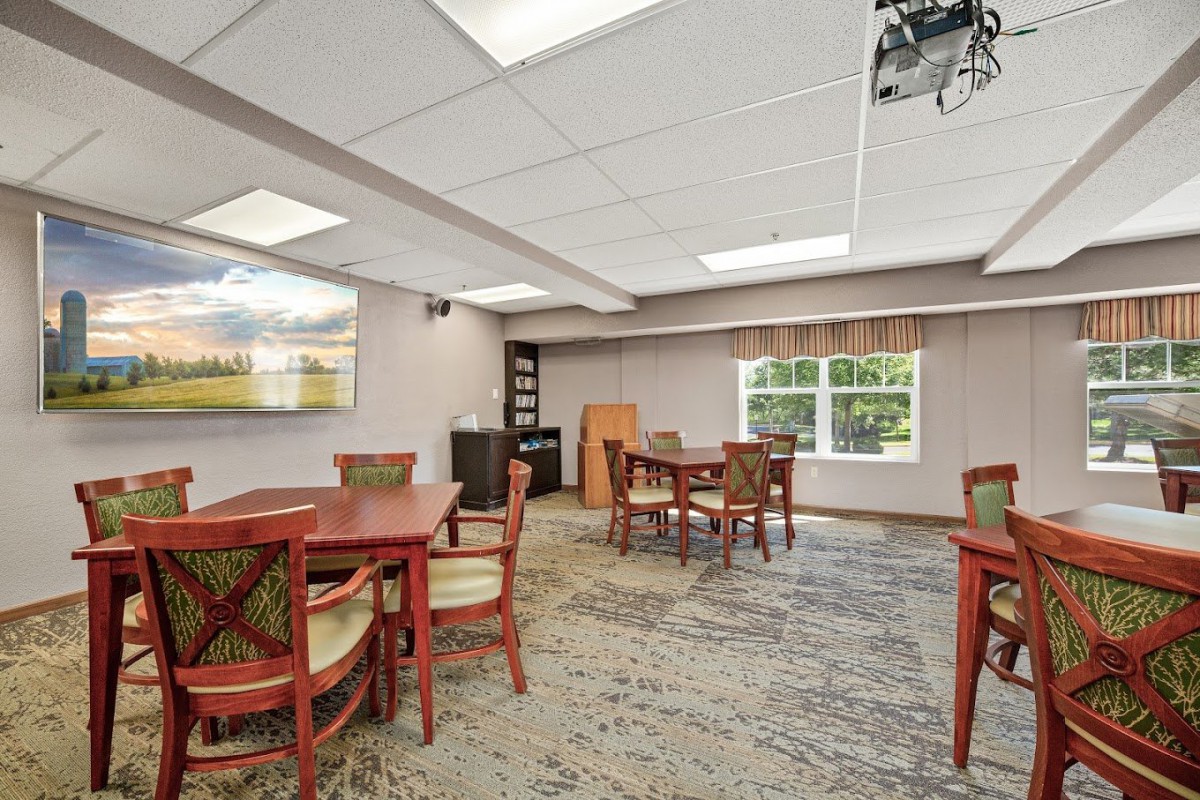 A well-lit conference room featuring multiple tables and chairs, a bookshelf, and a projector.