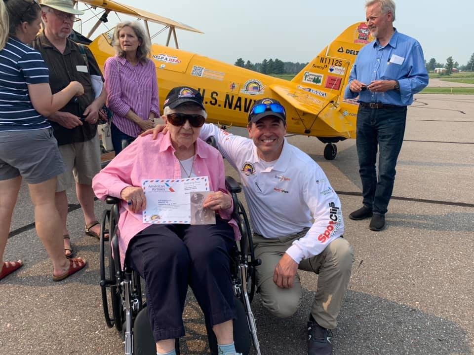 A group of people gather around a yellow U.S. Navy airplane. One person in a wheelchair holds a certificate, while another kneels beside them, smiling.