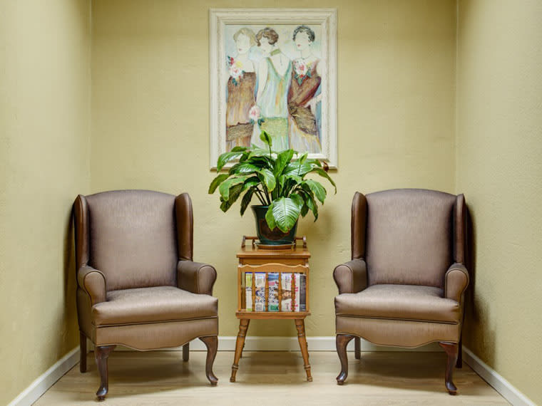 A cozy waiting area featuring two armchairs, a potted plant on a side table, and artwork on the wall.