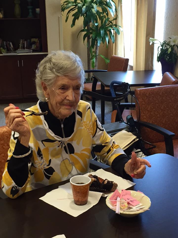 An elderly woman enjoys dessert and coffee while sitting at a table indoors.