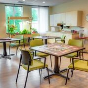 A cozy cafeteria with tables and chairs, featuring a kitchenette and indoor plants.