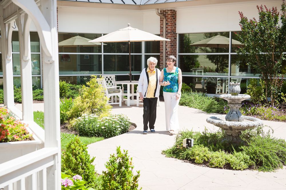 An elderly woman is walking with a caregiver in a sunny garden patio area.