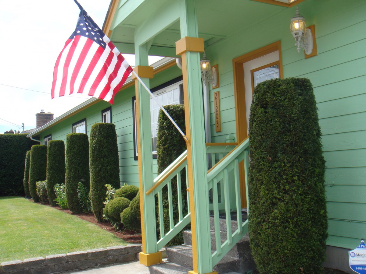 A green house with a porch and an American flag displayed, surrounded by neatly trimmed bushes.