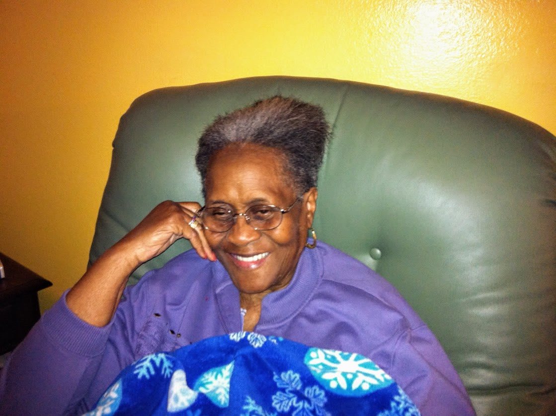 An elderly woman wearing glasses and a purple sweater, sitting comfortably in a chair with a blanket.