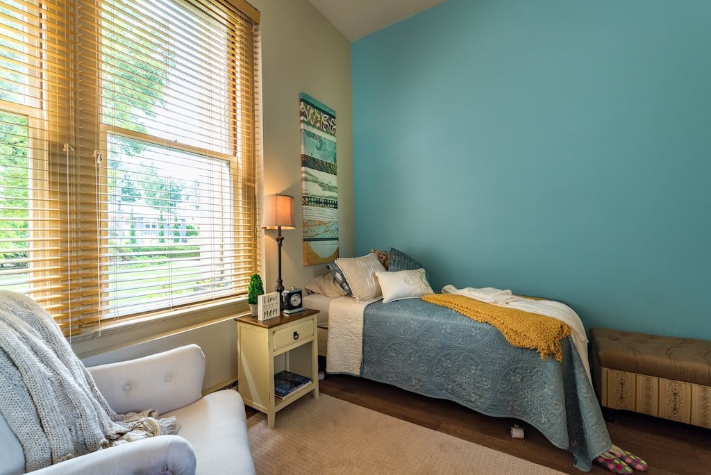 A cozy bedroom with a twin bed, featuring a blue accent wall and natural light through blinds.