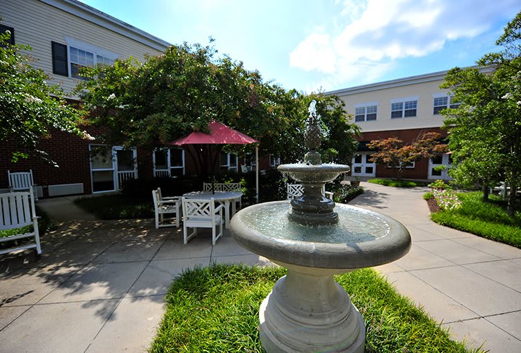 A tranquil courtyard featuring a fountain, outdoor seating with tables and chairs, shaded by umbrellas, surrounded by greenery and adjacent to a building.
