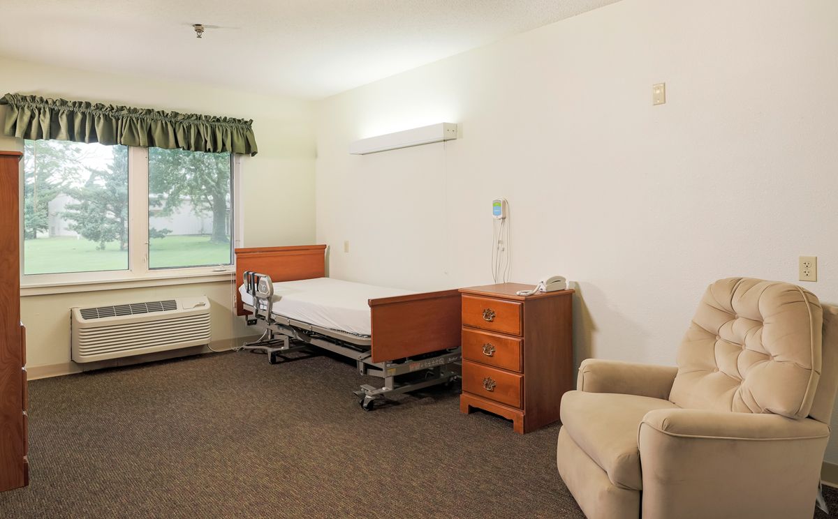 A minimalist hospital room with a single bed, dresser, and chair.